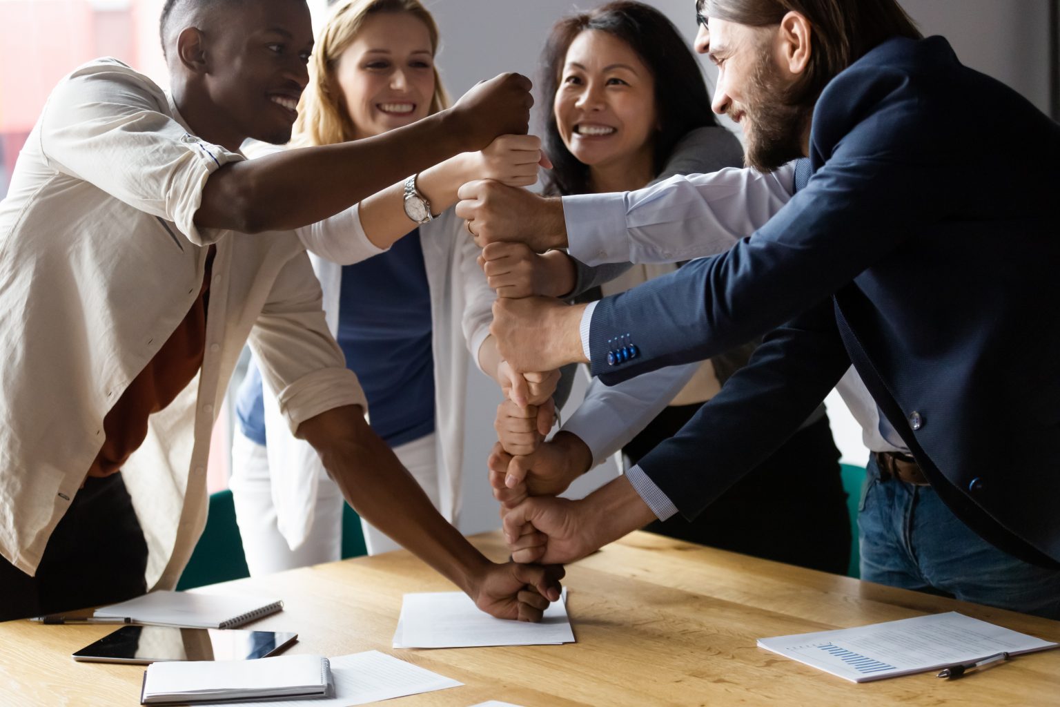 Happy diverse employees stack fists at corporate meeting, celebrating success, business achievement, engaged in funny team building activity, smiling colleagues showing unity and support